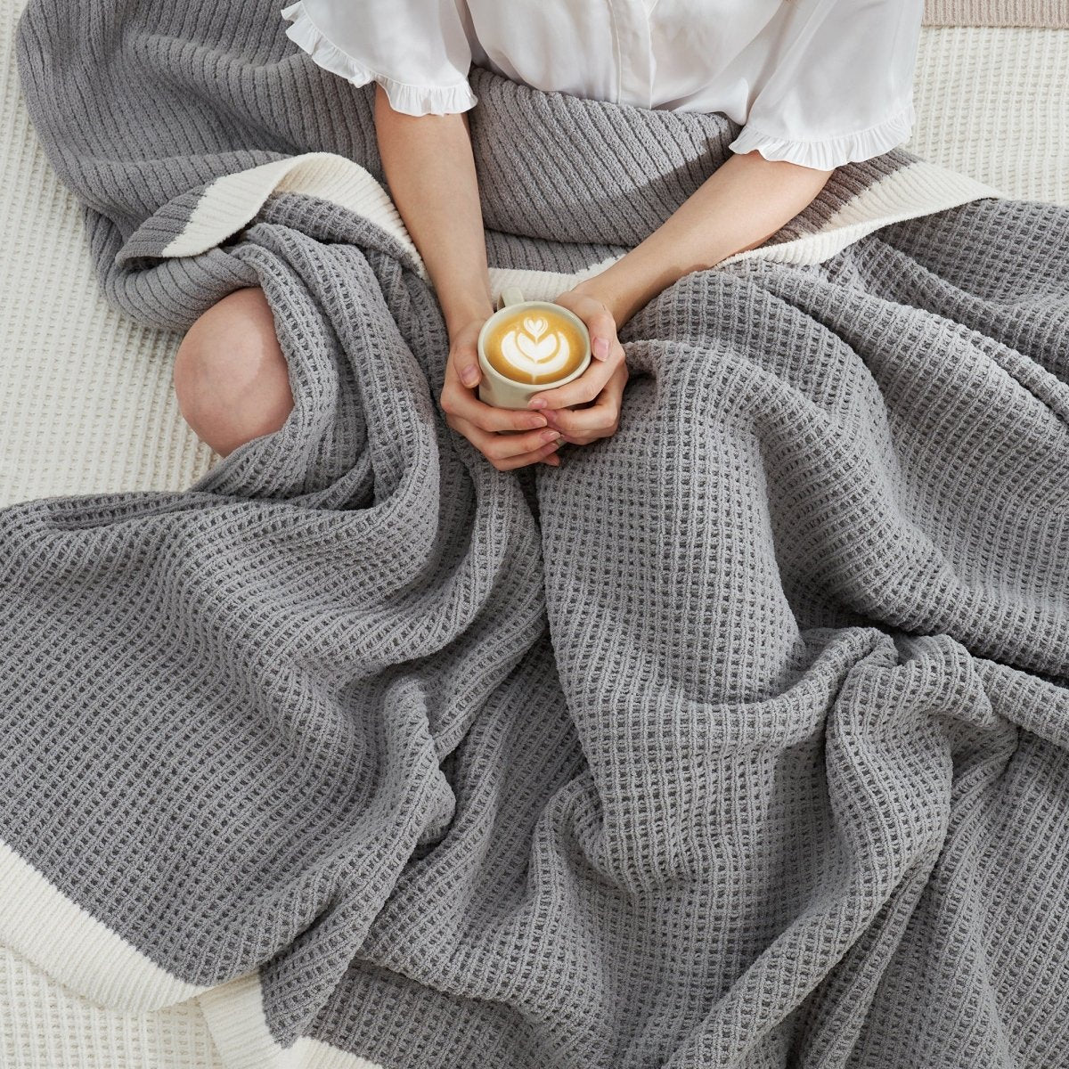 Woman wrapped in a Chenille Waffle Knit Throw Blanket in Grey, enjoying a cup of coffee on a sofa. #color_Grey