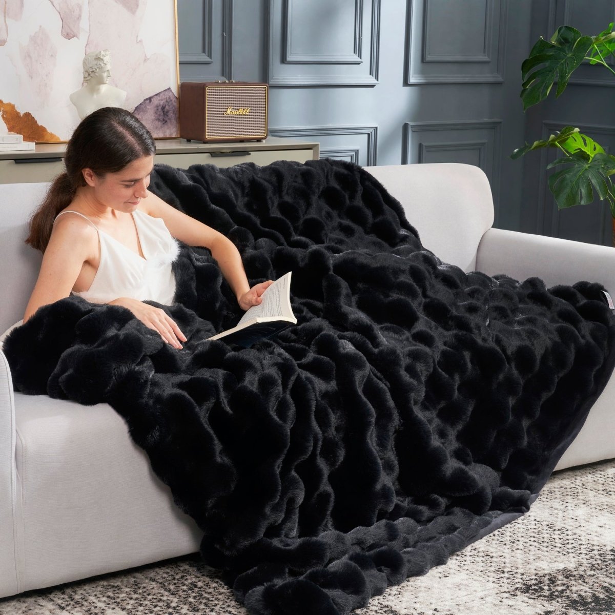 Women reading a magazine while wearing Cozy Bliss black bubble faux fur throw blanket on a couch. #color_Black