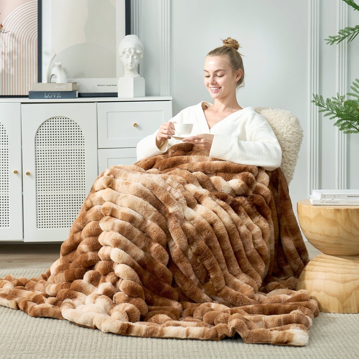 Woman sitting on a couch wrapped in a large brown fuzzy blanket, holding a cup. #Color_Tie-dye Brown