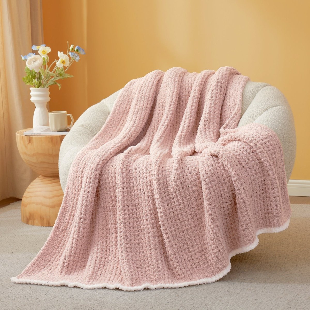 Pink textured blanket draped over a chair with a side table and flowers in the background. #color_Pink