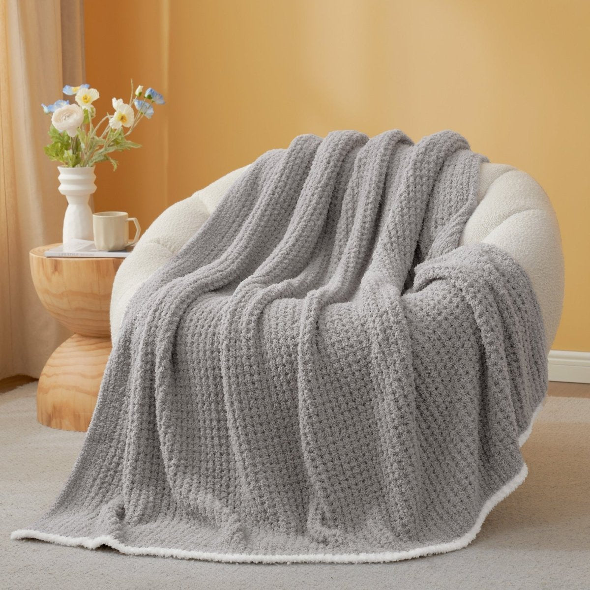 Grey textured blanket draped over a chair with a side table and flowers in the background. #color_Grey