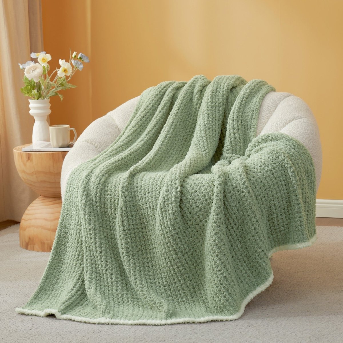 Sage Green textured blanket draped over a chair with a side table and flowers in the background. #color_Sage Green