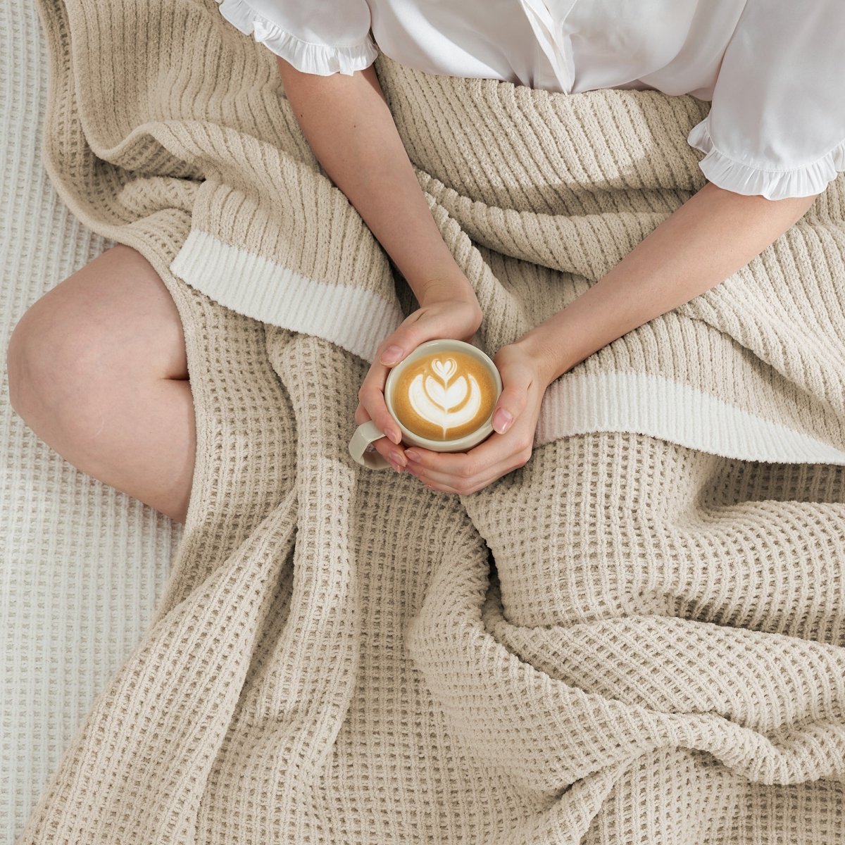 Woman wrapped in a Chenille Waffle Knit Throw Blanket in beige, enjoying a cup of coffee on a sofa. #color_Beige