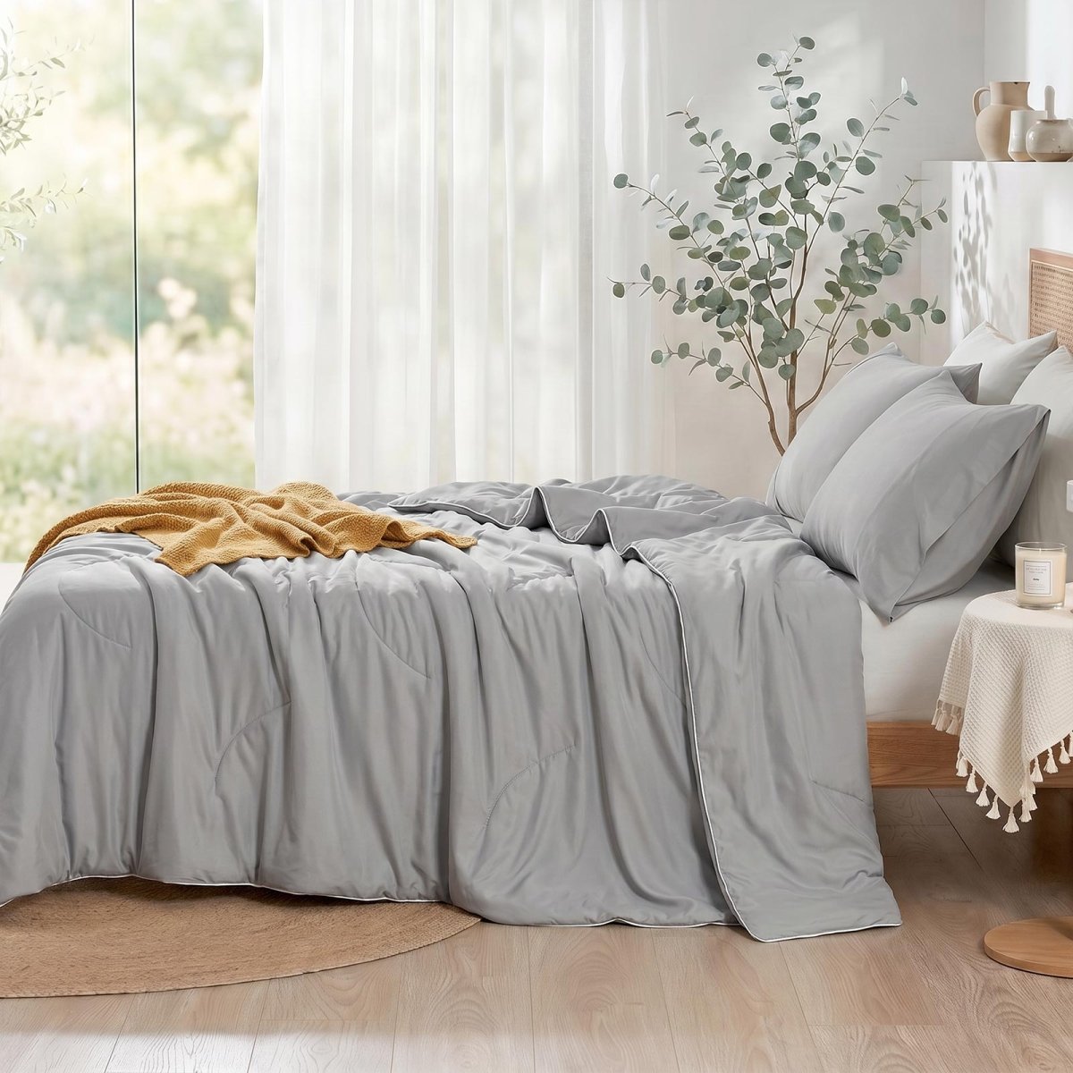 Cozy Bliss double-sided cooling comforter on a bed in a bright, sunlit bedroom with green plants, lightweight summer bedding for hot sleepers.#color_Cool Grey