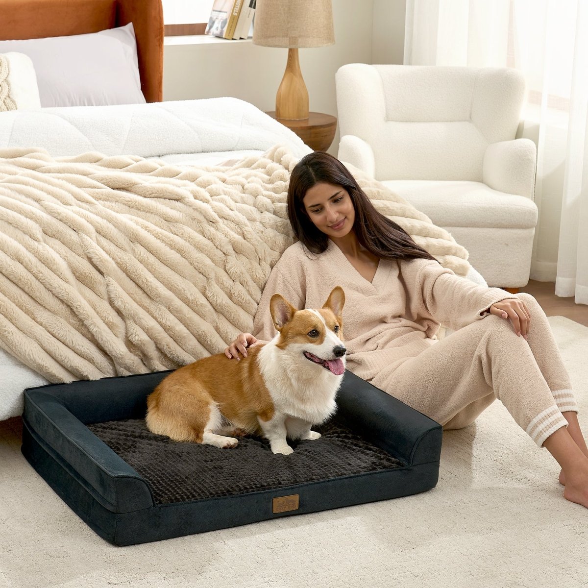 Dog lying on a black pet bed in a living room with a woman sitting beside it. #color_Black